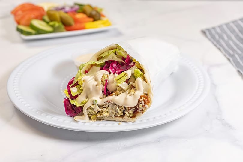 A falafel wrap with lettuce, red cabbage, and tahini sauce on a white plate, with a side salad.